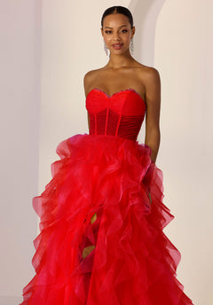 A woman in the Morilee 49218 long prom dress in poppy, featuring a strapless corset bodice and voluminous ruffled organza skirt, stands indoors against a light background.