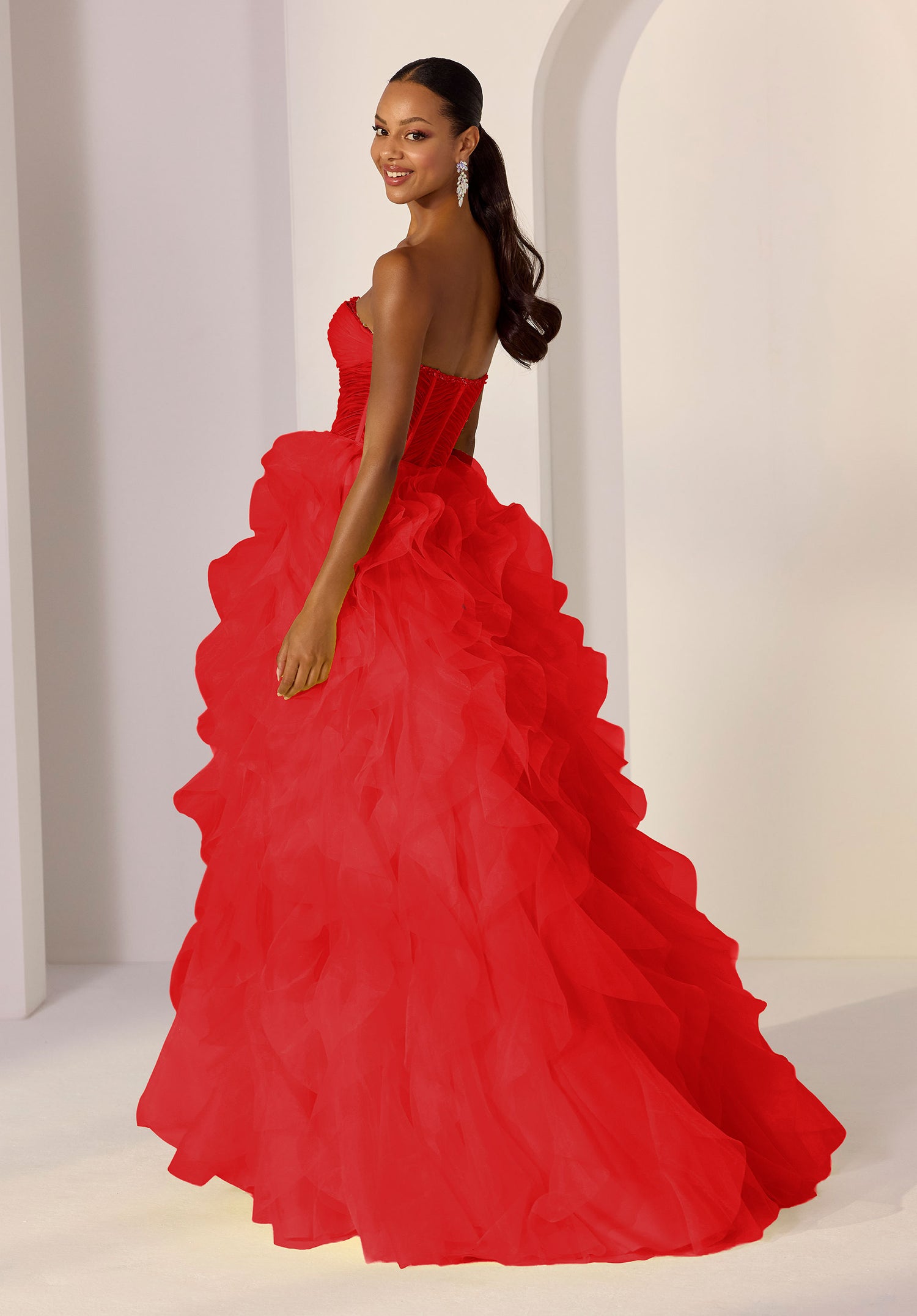 A woman in a bright red, ruffled Morilee 49218 long prom dress stands on a white floor, looking over her shoulder with a neutral expression.