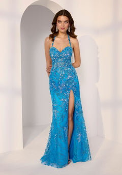 A woman in a turquoise Morilee 49243 long prom dress with lace-applique halter straps, silver embroidery, and a high slit stands before a white arched backdrop.