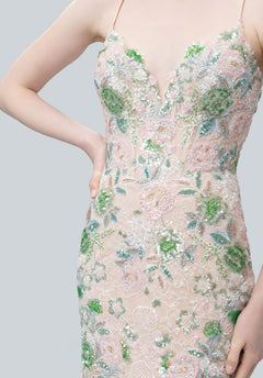 Someone wearing the Morilee 49244 Long Prom Dress, featuring a sleeveless V-neck and green, pink, and white floral embroidery on sheer fabric, stands with one hand on their hip.