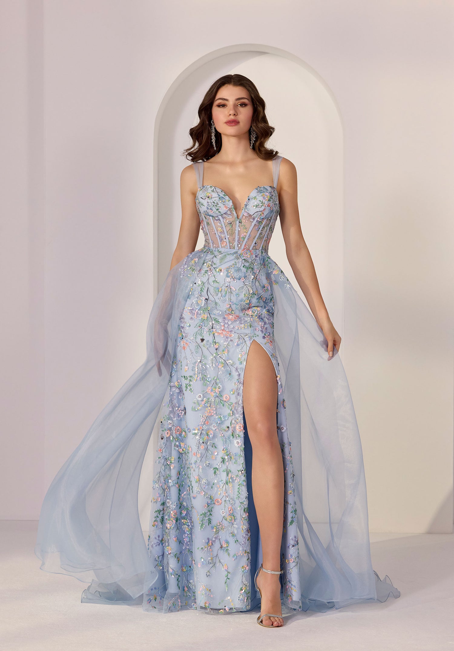 A woman in the Morilee 49249 long prom dress, featuring light blue floral sequins, a sheer corset bodice, thigh-high slit, and tulle overlay, stands before a white arched background.