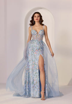 A woman in the Morilee 49249 long prom dress, featuring light blue floral sequins, a sheer corset bodice, thigh-high slit, and tulle overlay, stands before a white arched background.