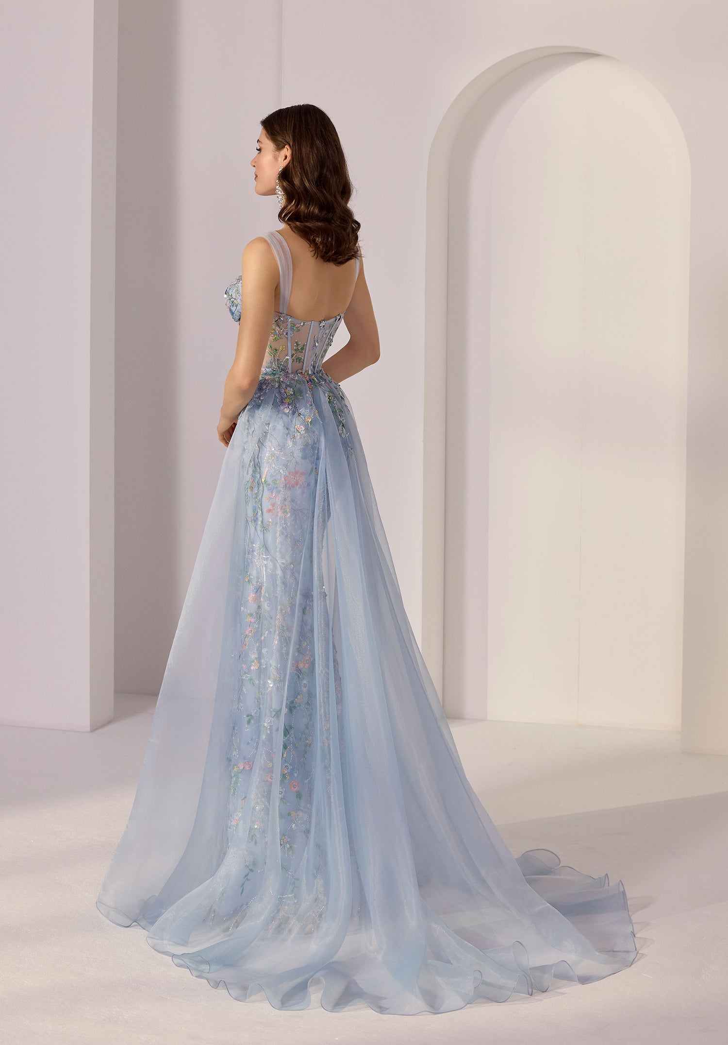 A woman stands indoors near an arched doorway, facing away from the camera in the Morilee 49249 long prom dress—a light blue floral sequin gown with sheer details.