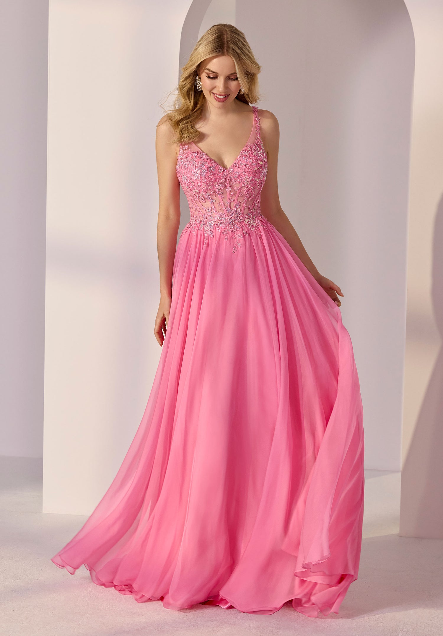 A woman wears the Morilee 49250 Long Prom Dress in fuchsia, featuring a flowing, floor-length skirt, fitted embellished bodice with delicate floral appliqué and thin straps, posed before a light background with arched architecture.