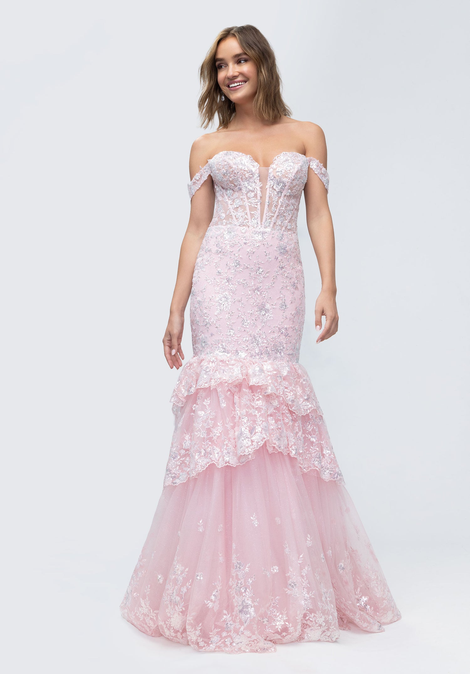 A woman in the Morilee 49274 Long Prom Dress—an off-shoulder, light pink mermaid gown with lace corset, floral accents, and a tiered tulle skirt—stands against a plain background, smiling.