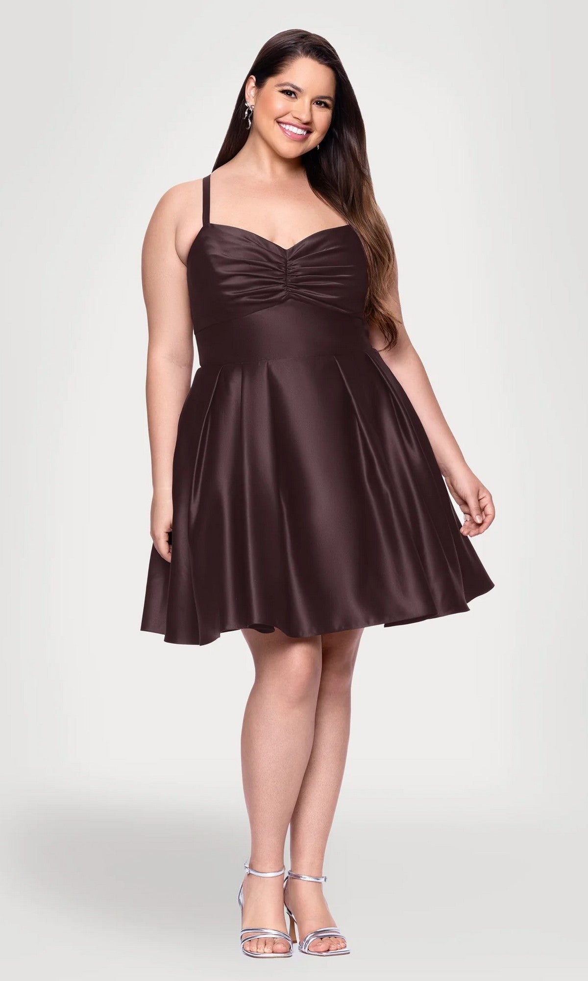 A woman wearing the Plus Short Wine Hoco Dress, Blondie Nites 4927BNW—a sleeveless, knee-length plus-size cocktail dress with thin straps—pairs it with silver heels and stands against a plain light gray background.