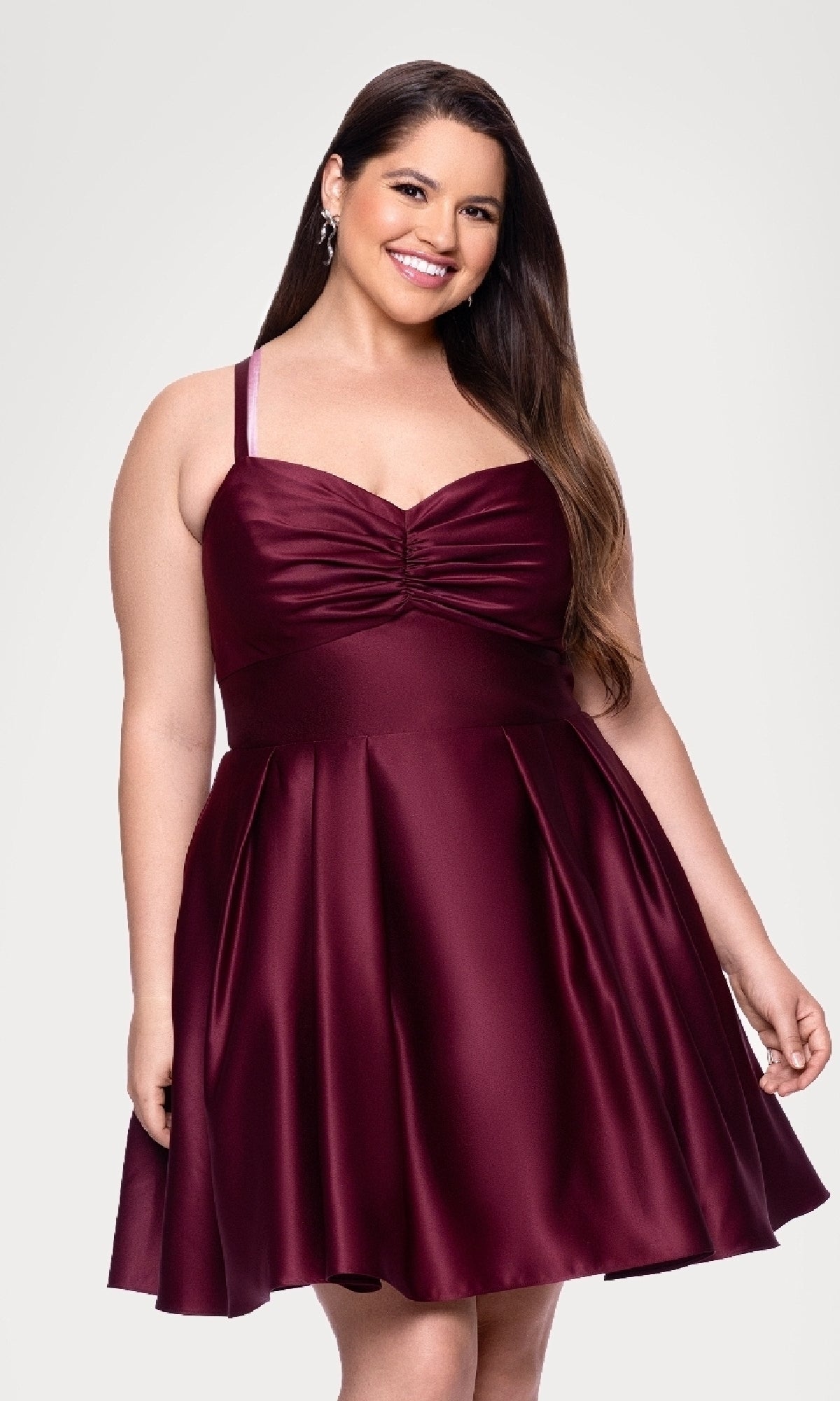 A woman smiles at the camera in the Plus Short Wine Hoco Dress Blondie Nites 4927BNW, a sleeveless, knee-length wine-colored party dress with a ruched bodice, standing against a plain light background.