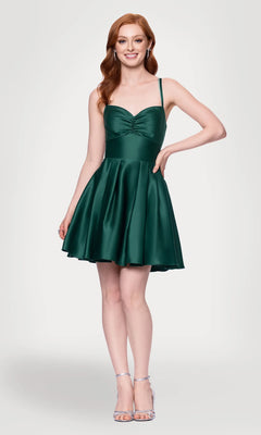 A woman with long red hair wears the Hunter Green Mini Hoco Dress: Blondie Nites 4927BN, a short green dress with thin straps, paired with silver high-heeled sandals, standing against a plain light background.