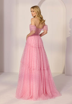 A woman stands in a bright room wearing the Morilee 49281—a floor-length pink off-the-shoulder prom dress with layered tulle and textured polka dots—glancing over her shoulder.