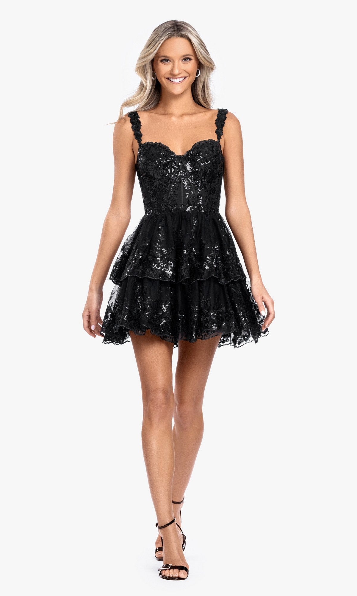 A woman in the Short Ruffled Hoco Dress: Blondie Nites 4931BN in black, featuring sequins, thin straps, and a tiered mini silhouette, stands against a white background wearing black strappy heels—perfect for homecoming.