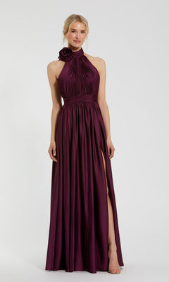 A woman wears the Mac Duggal 49333 high-neck halter formal dress—sleeveless, floor-length, in dark purple—with a pleated bodice, floral shoulder detail, and a high side slit.