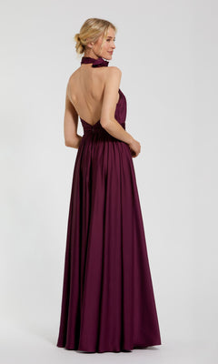 A woman in the Mac Duggal 49333 High-Neck Halter Formal Dress, a floor-length, sleeveless gown with an open back, stands slightly turned away against a plain light background.
