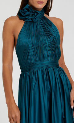 A woman wears the Mac Duggal 49333 High-Neck Halter Formal Dress—a sleeveless teal satin gown featuring pleated bodice detailing and a large fabric flower at the high neckline.