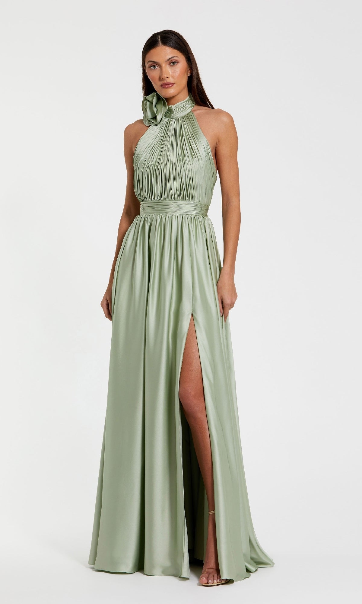 A woman stands against a plain white background wearing the Mac Duggal 49333 High-Neck Halter Formal Dress in light sage green, featuring a floor-length skirt, pleated bodice, high neckline, and thigh-high slit.