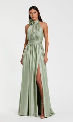 A woman stands against a plain white background wearing the Mac Duggal 49333 High-Neck Halter Formal Dress in light sage green, featuring a floor-length skirt, pleated bodice, high neckline, and thigh-high slit.