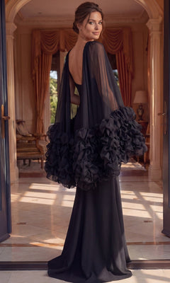 A woman in the Jovani 49448 long formal dress, featuring sheer ruffled sleeves and a sleek black designer silhouette, stands in an elegant room adorned with arched doorways and gold curtains, glancing over her shoulder.
