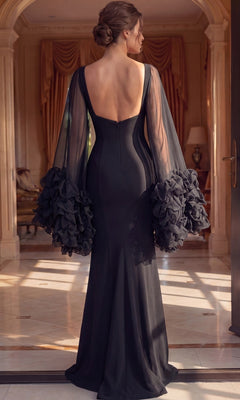 A woman wearing the Long Formal Dress: Jovani 49448—a black, floor-length gown featuring sheer, ruffled sleeves—stands indoors, facing away in a well-lit and elegant room.