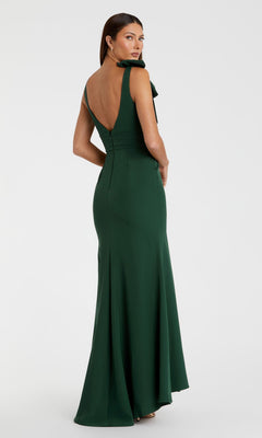 A woman stands looking over her shoulder in the Mac Duggal 49454 Shoulder-Bow Long Prom Dress, a floor-length dark green gown featuring a deep V-back and elegant bow detail on the shoulder.