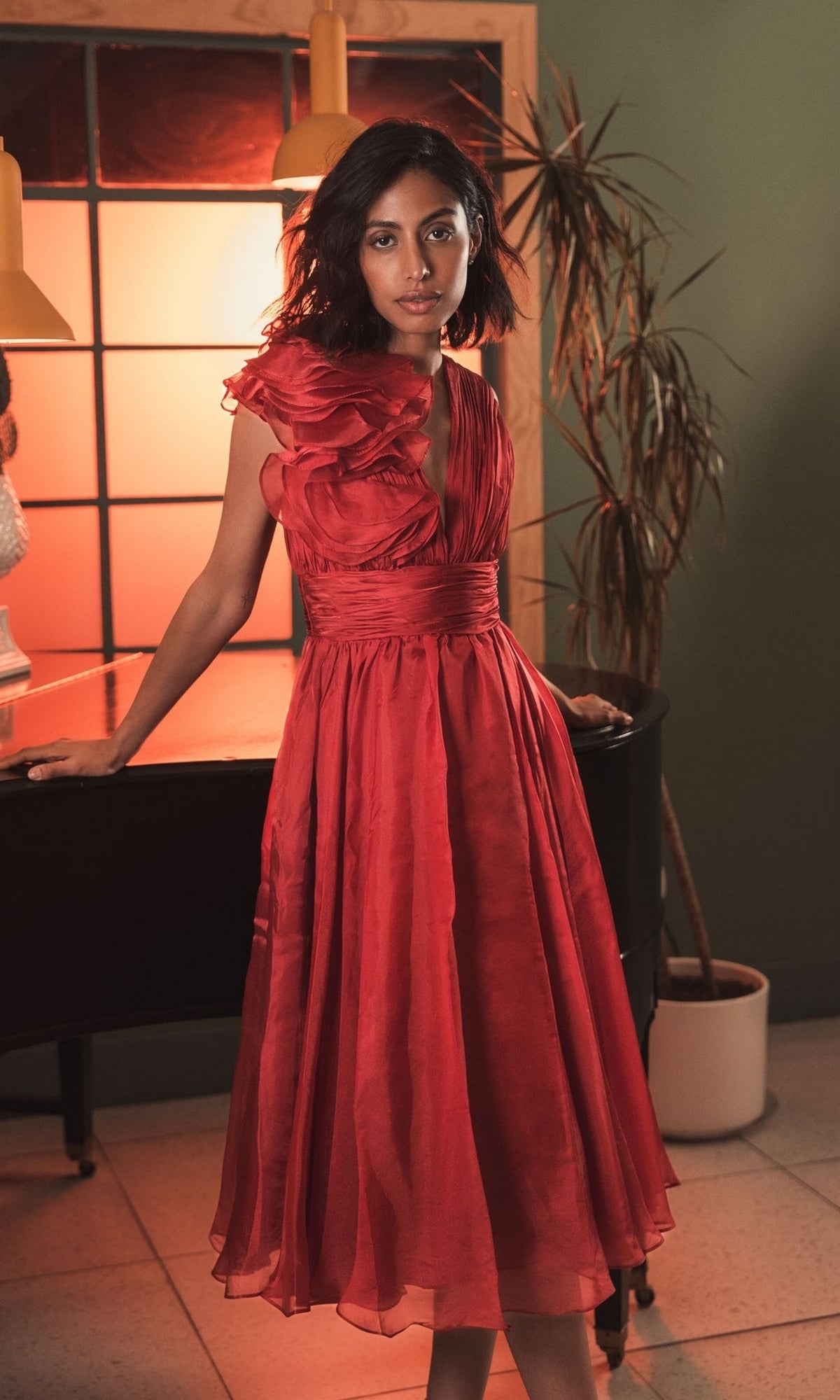 A woman stands indoors by a piano and tall plant, illuminated by warm light and a window, wearing the Mac Duggal 49494 short ruched wedding-guest dress.