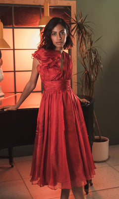 A woman stands indoors by a piano and tall plant, illuminated by warm light and a window, wearing the Mac Duggal 49494 short ruched wedding-guest dress.