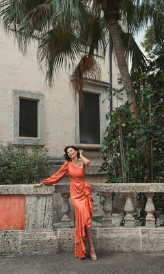 A woman wearing the Long Formal Dress 49531 by Mac Duggal, an orange one-shoulder gown, smiles and touches her hair outdoors by a stone balustrade, with greenery and a stucco building in the background.