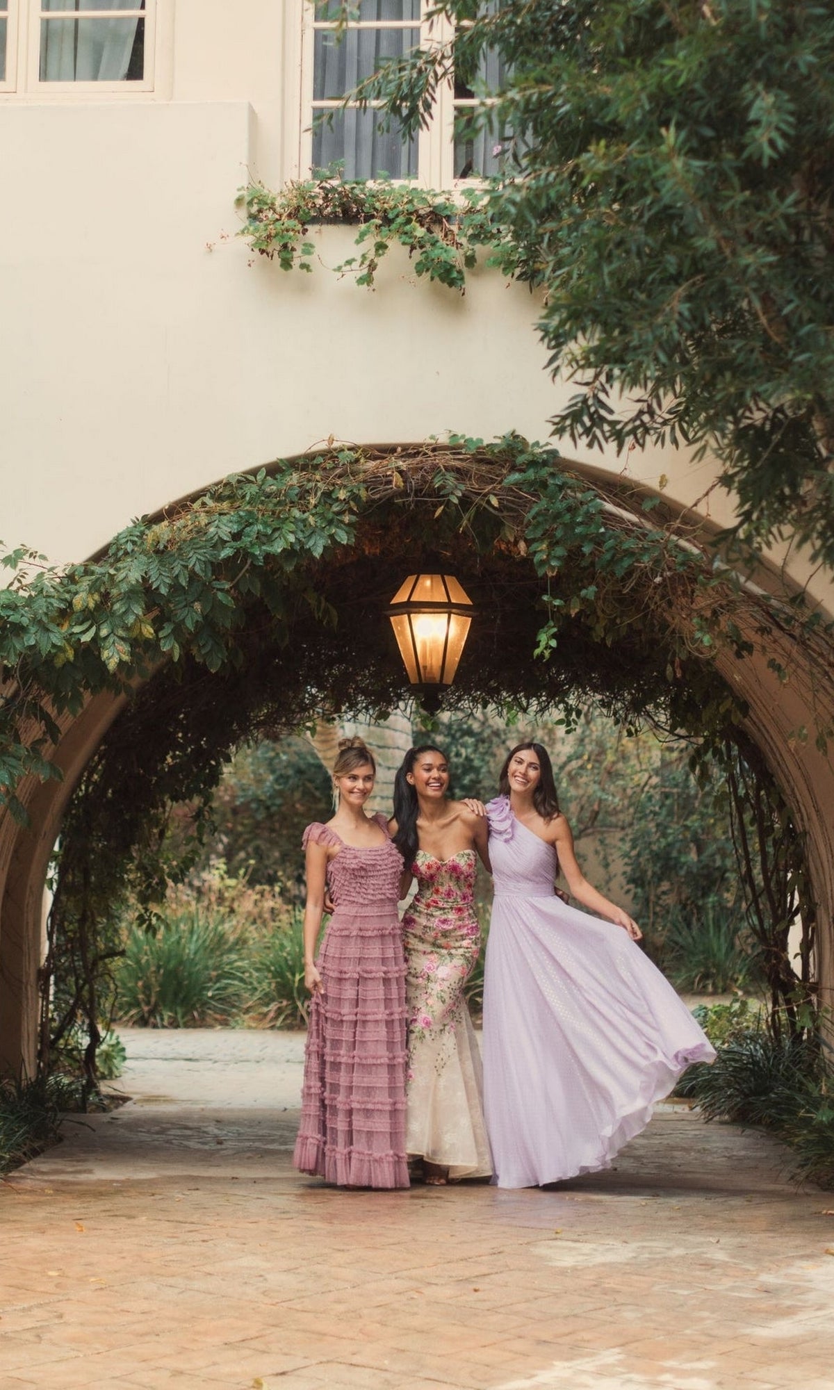 Three women wearing the Mac Duggal 49536 One-Shoulder Long Formal Dress pose together, smiling under an ivy-covered archway on a stone path.