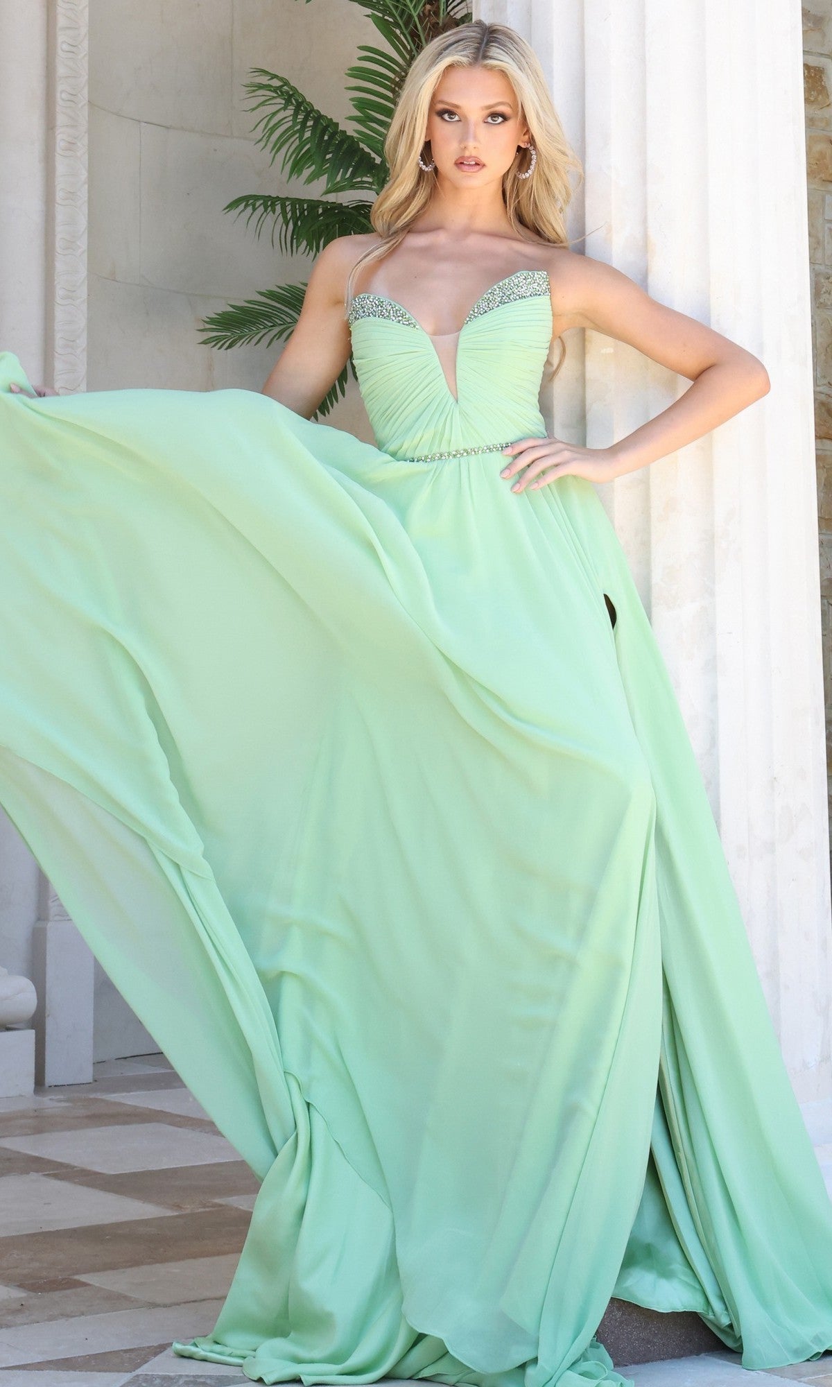 The woman stands in front of white columns and a green plant, wearing the Jovani 49536 long formal dress in mint—a strapless chiffon gown featuring a flowing skirt and subtle crystal-embellished trim.