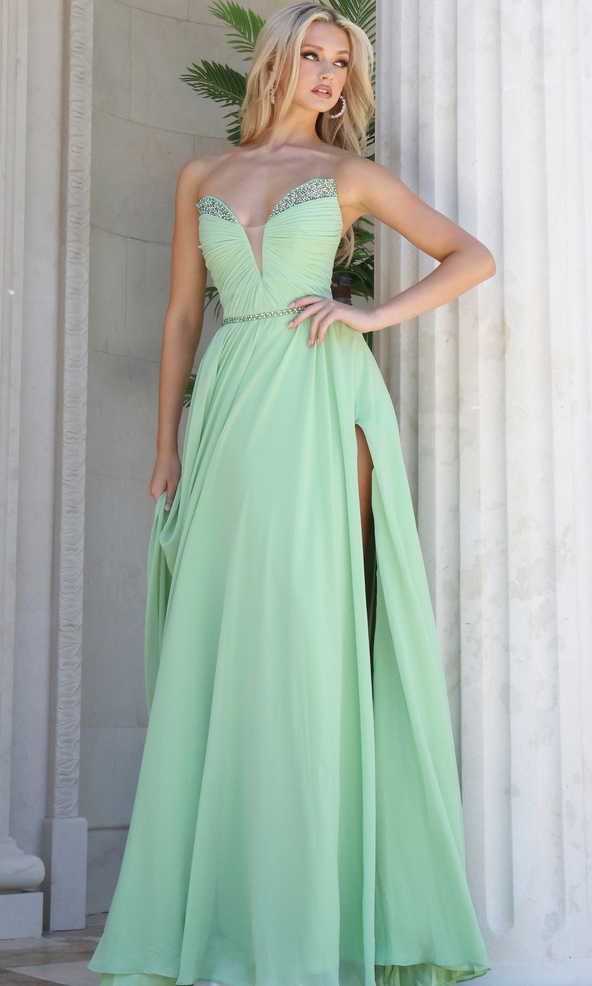 A woman wearing the Jovani 49536 long formal dress in light green chiffon, featuring a sweetheart neckline and side slit, stands in front of white columns and greenery.