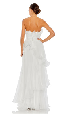 A woman stands facing away, wearing the Long Formal Dress 49537 by Mac Duggal—a strapless white gown with ruffles, a pleated floor-length skirt, and a subtle empire waist. Her hair is styled in a low bun.