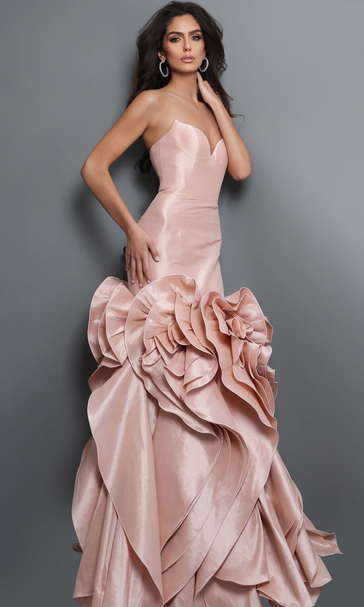 A woman wearing the Jovani 49548 long formal dress in blush, featuring a strapless neckline and large, sculpted ruffles on the lower half, stands against a plain gray background.