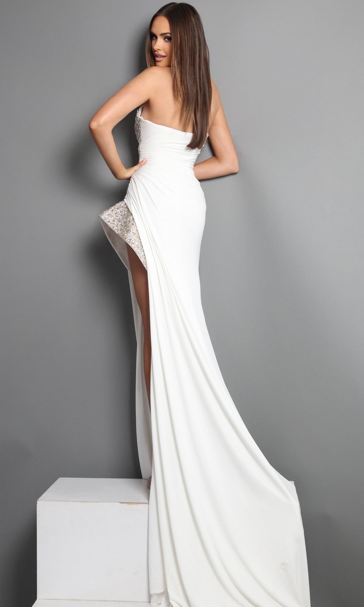 A woman poses on a platform in the Jovani 49577, a long white strapless crepe formal dress, showing off its column silhouette and high slit as she looks over her shoulder.