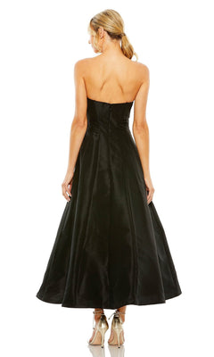 A blonde woman in a low ponytail wears the strapless black Mac Duggal 49605 tea-length wedding-guest dress and metallic heels, standing with her back to the camera against a white background.