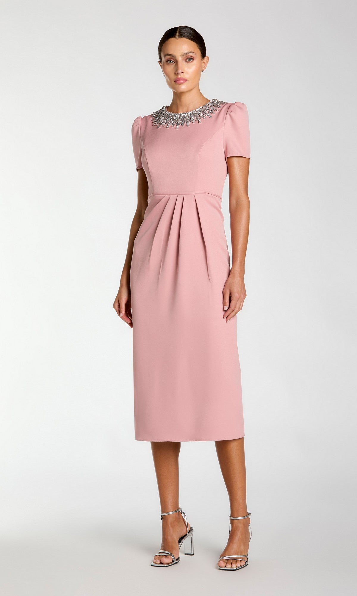 A woman wears the Mac Duggal 49606 short sleeve tea-length party dress in light pink, featuring a beaded collar and front pleats, paired with silver heels. She poses against a plain light background.