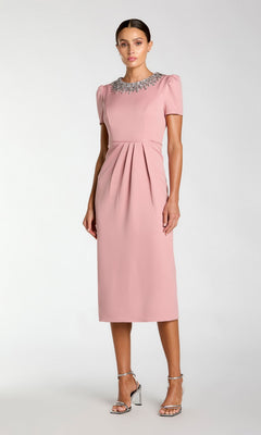 A woman wears the Mac Duggal 49606 short sleeve tea-length party dress in light pink, featuring a beaded collar and front pleats, paired with silver heels. She poses against a plain light background.
