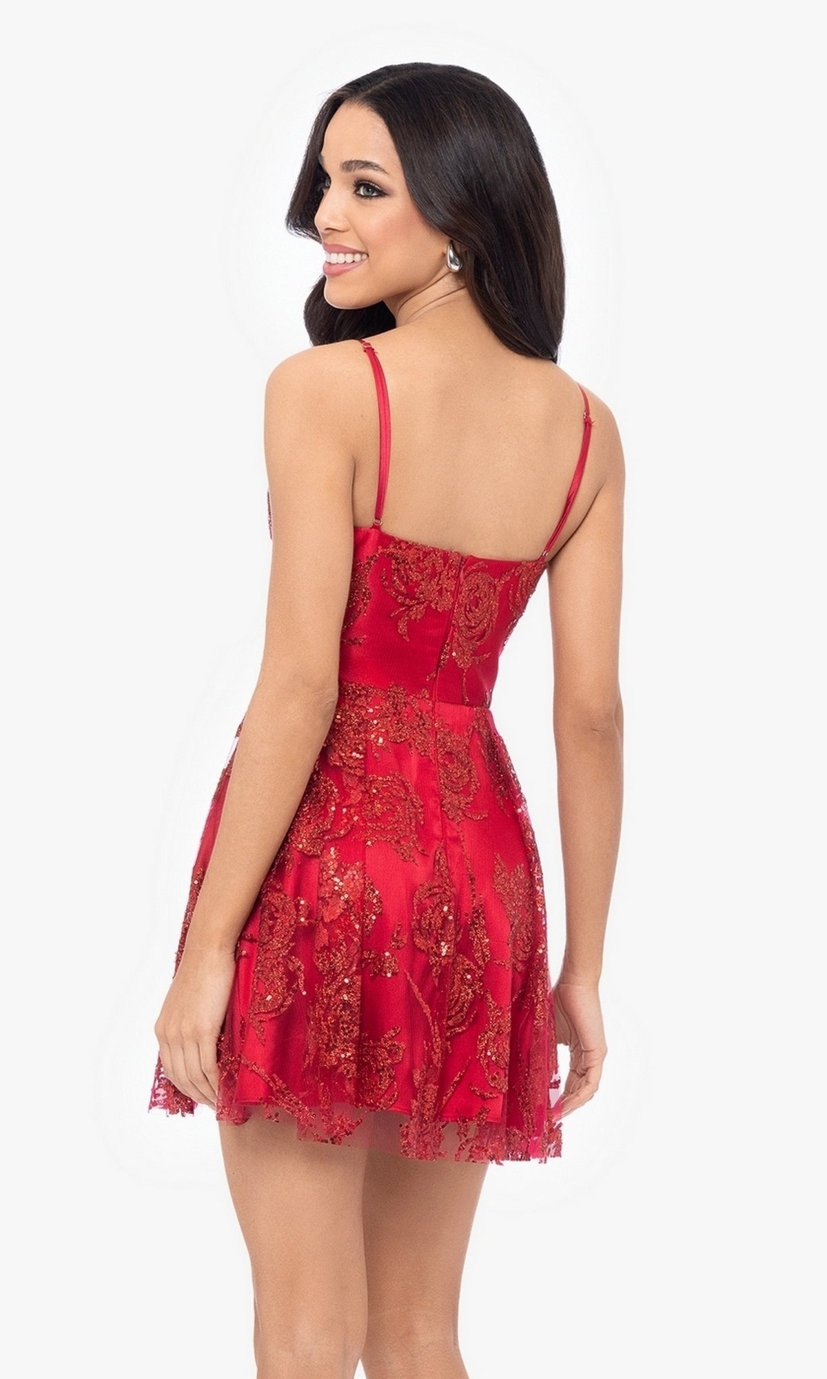 A woman wearing the Short Red Glitter Homecoming Dress 4960BN—a sleeveless, short red dress with glitter detail—stands facing away, smiling over her shoulder.