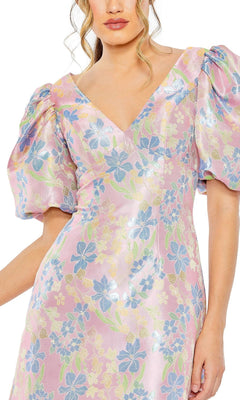 A woman wears the Short Homecoming Dress 49622 by Mac Duggal—a shiny pink dress with blue and yellow floral patterns and puffed short sleeves.