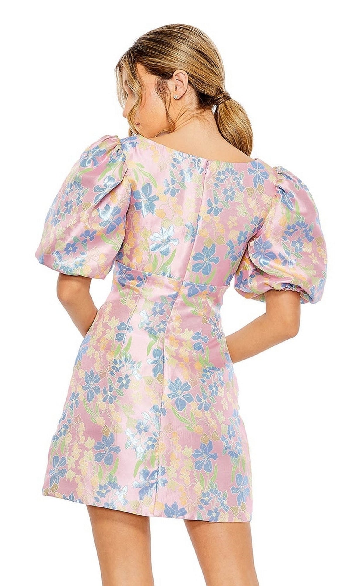 A woman with light brown hair in a low ponytail is seen from the back wearing the Mac Duggal Short Homecoming Dress 49622, featuring puffed sleeves and a pastel floral print.