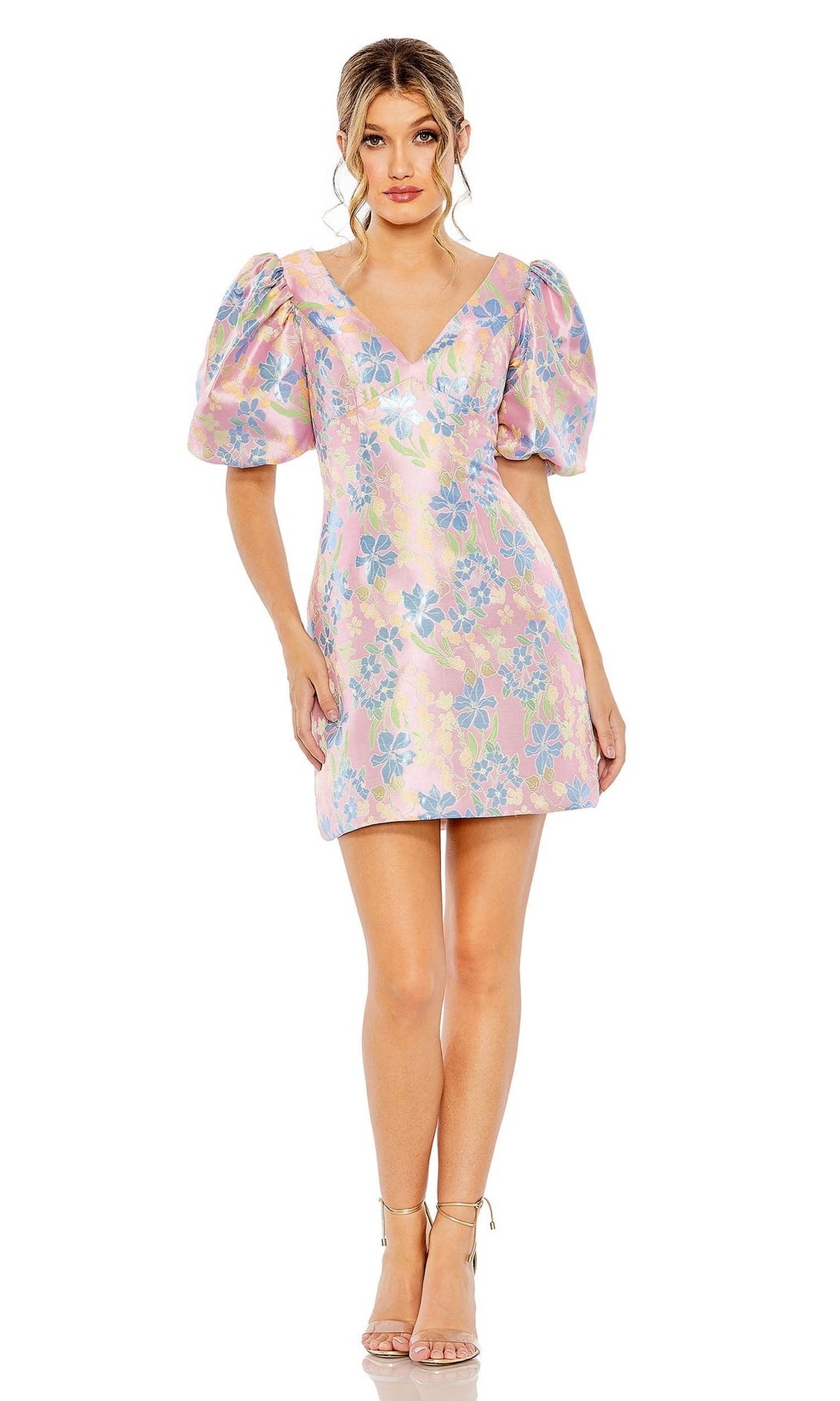 A woman wears the Short Homecoming Dress 49622 by Mac Duggal, a pastel floral print dress with puffed sleeves and a V-neckline, paired with nude high-heeled sandals against a plain white background.