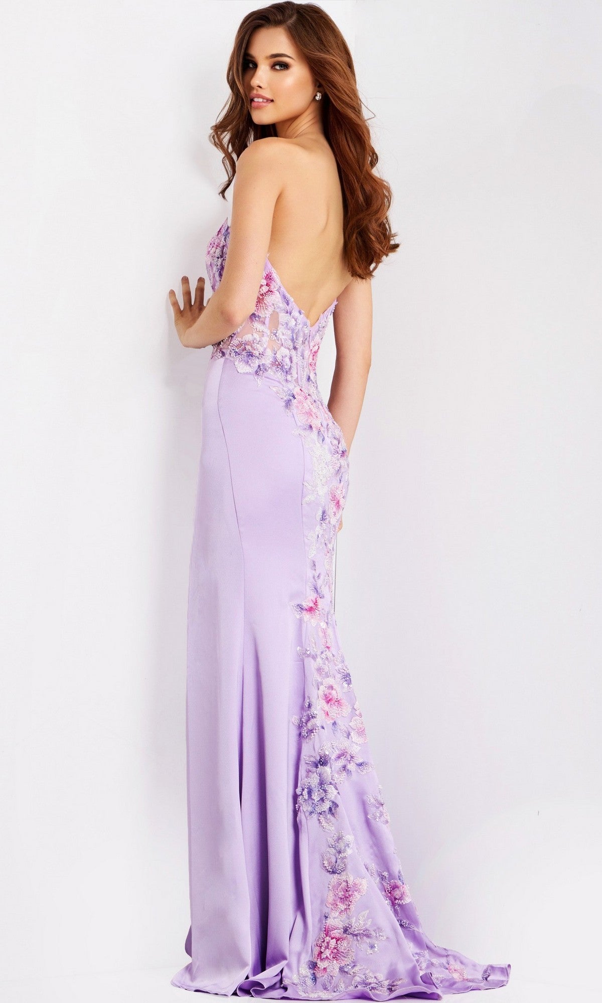 A woman models the Jovani 49629 long prom dress—a floor-length, light purple sheath gown with floral embroidered appliques—standing and glancing over her shoulder against a plain background.