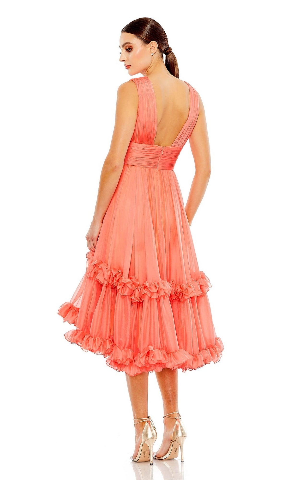 A woman in the Mac Duggal Short Wedding Guest Dress 49635—a sleeveless, ruched peach knee-length dress with ruffled layers and a fitted waistband—stands in high heels against a white background.