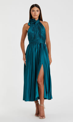 A woman in the Mac Duggal 49674 Halter Midi Wedding Guest Dress—teal, sleeveless with a floral shoulder detail, pleated bodice, and thigh-high slit—creates an elegant semi-formal look paired with strappy heels.