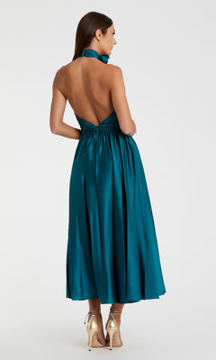 A woman in the Mac Duggal 49674 ocean blue halter midi wedding guest dress with an open back and gold high heels stands with her back to the camera against a plain background.