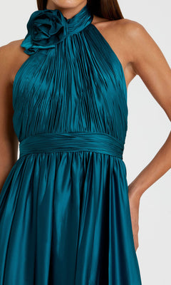 A woman wears the Halter Midi Wedding Guest Dress: Mac Duggal 49674—an ocean blue, sleeveless, pleated satin dress with a fitted waistband and large neckline flower. A chic semi-formal choice for special occasions.
