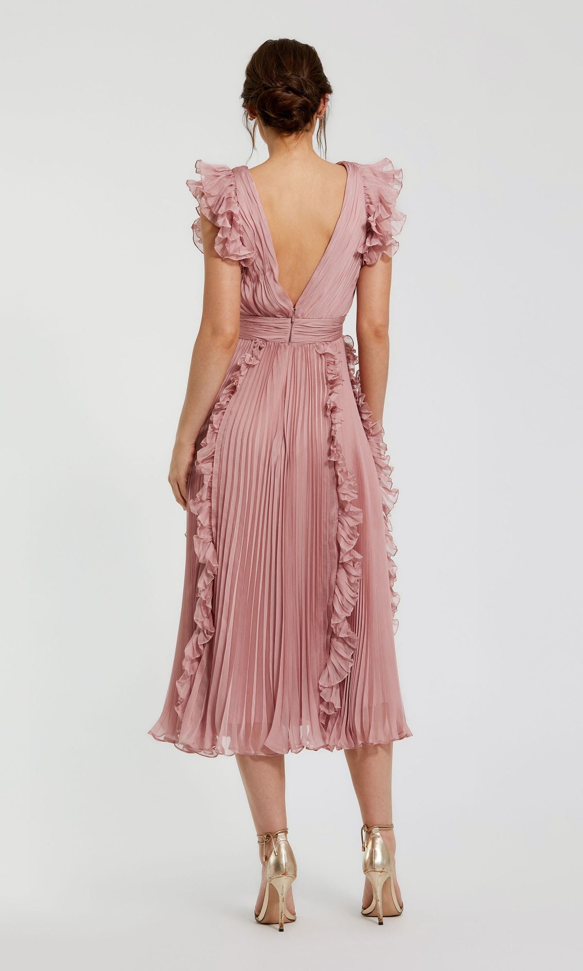 A woman stands with her back to the camera in the rose pink Short Wedding Guest Dress 49675 by Mac Duggal, featuring chiffon fabric, ruffle cap sleeves, a pleated hem, and paired with gold high-heeled shoes.