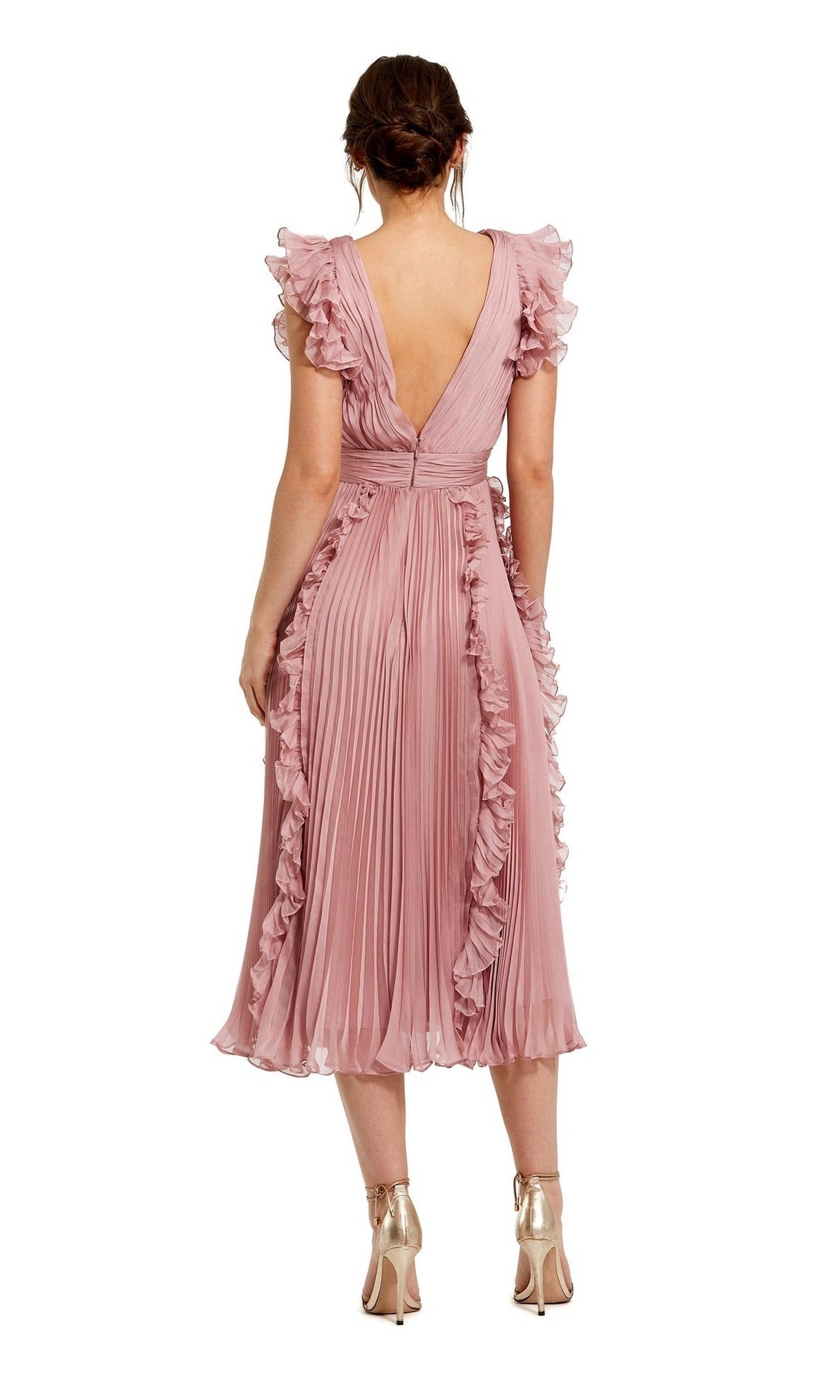 A woman stands facing away, wearing the Short Wedding Guest Dress 49675 by Mac Duggal—a rose pink chiffon midi dress with ruffle cap sleeves, pleated details—and metallic high-heeled shoes.