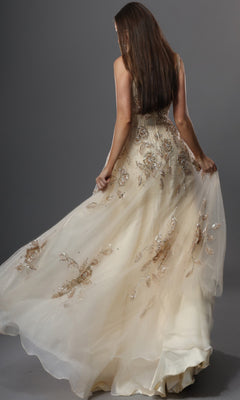 A woman with long brown hair is shown from the back, holding out the skirt of her embellished cream gown—Long Formal Dress: Jovani 49697—in front of a plain gray backdrop.