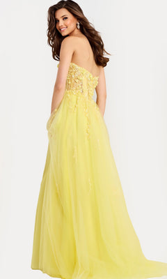 A woman models the Jovani 49704, a long strapless prom dress with a floral-embellished bodice, standing against a plain light background and smiling over her shoulder.