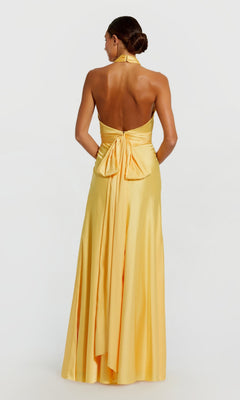 A woman faces away, wearing the Mac Duggal 49728 long formal dress—yellow satin with a halter neck, open back, and a large bow detail at the waist.