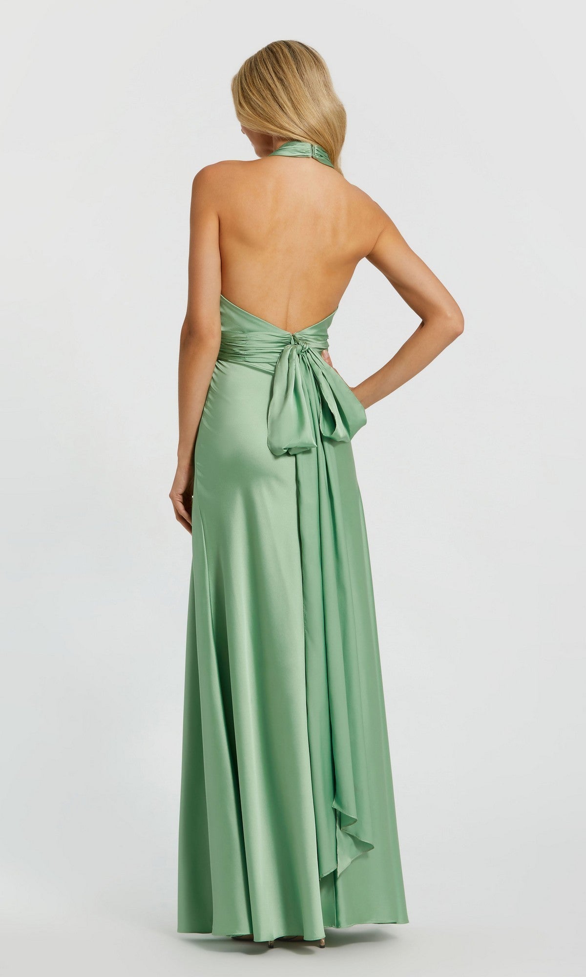 A woman stands facing away in the Mac Duggal 49728 long formal dress—a light green, halter-neck satin gown featuring a backless design, large bow, and flowing sash detail.
