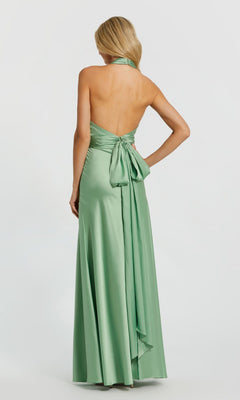 A woman stands facing away in the Mac Duggal 49728 long formal dress—a light green, halter-neck satin gown featuring a backless design, large bow, and flowing sash detail.